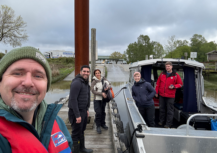 Team stands at and in an aluminum boat on an estuary near Vancouver smiling at the camera