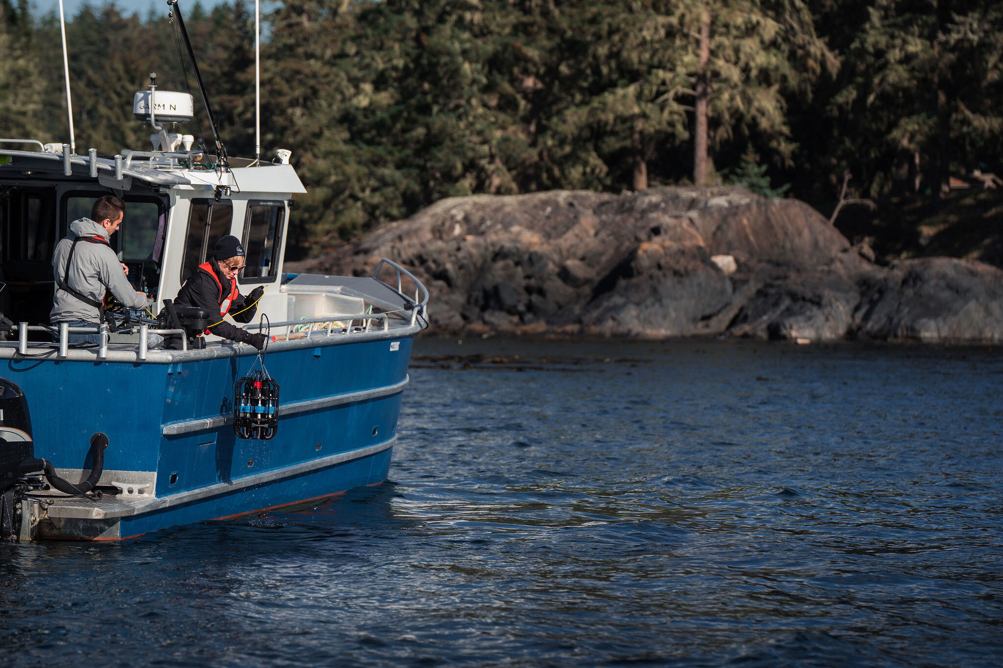 Two researchers stand at the side of a small aluminum boat. One lowers the Ascension depth sampler over the side by hand. Two researchers stand at the side of a small aluminum boat. One lowers the Ascension depth sampler over the side by hand.