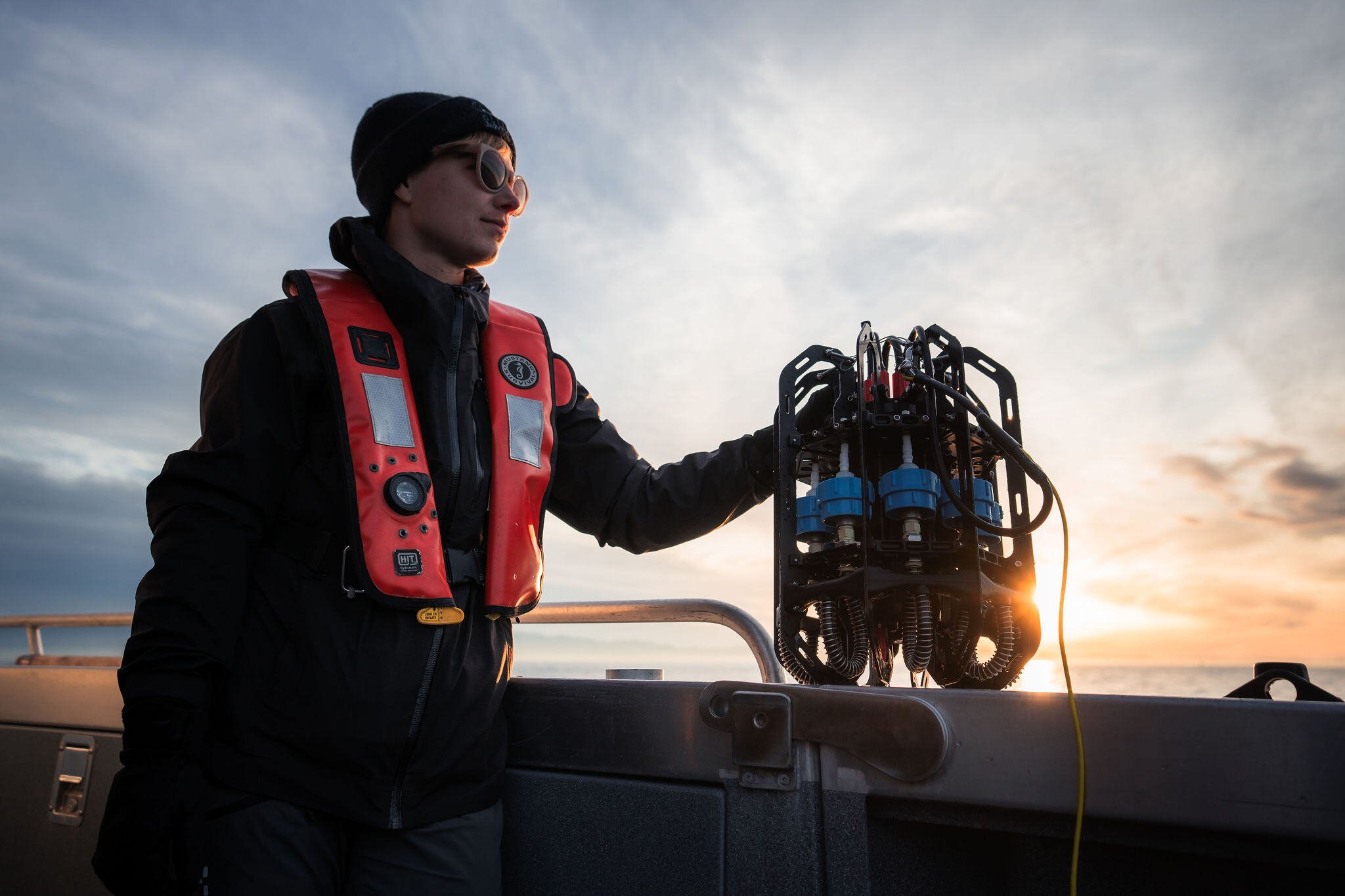 Scientist wearing a life vest stands at the rail of an aluminum boat during sunset with one hand on the Ascension eDNA depth sampler that sits upon the boat side.