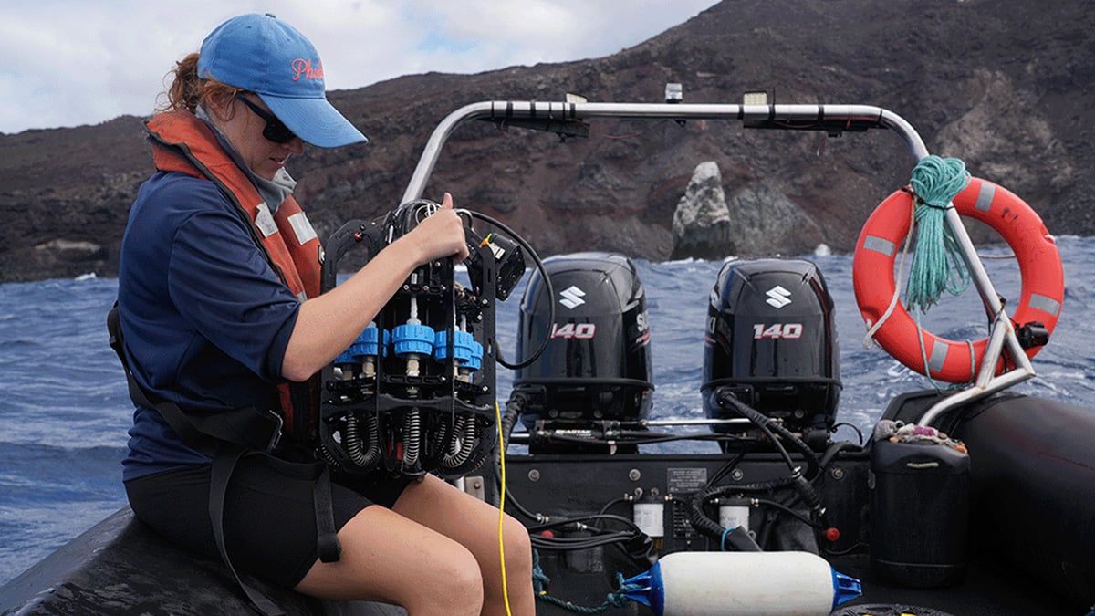 A scientists sits on the side of a small dinghy holding the Ascension eDNA depth sampler on her knee