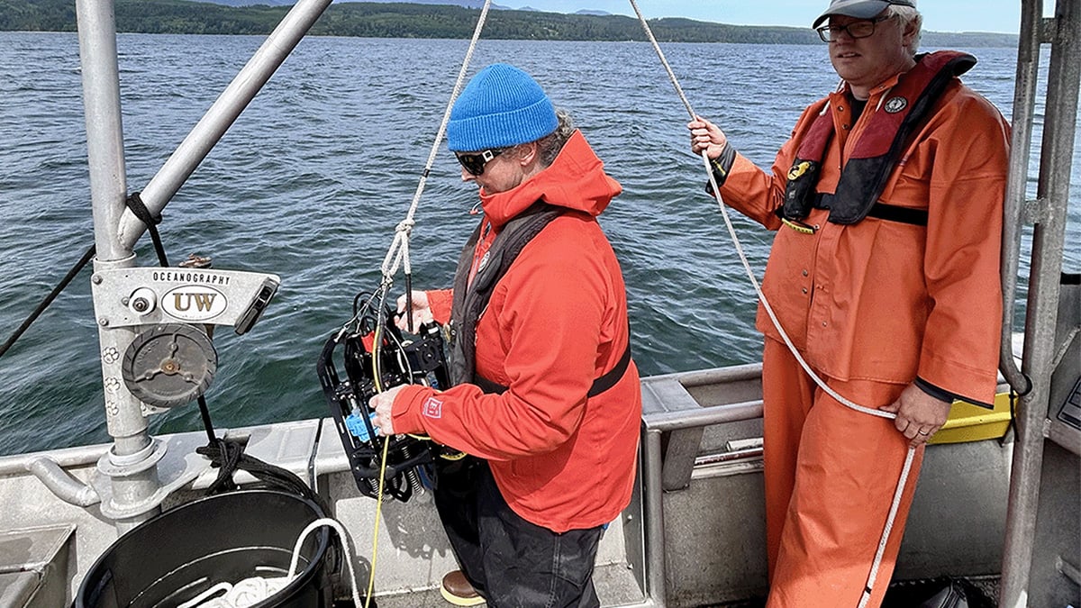 A woman researcher stands holding an Ascension eDNA depth sampler attached to a pull system held by another researcher on the side of a small aluminum fishing boat