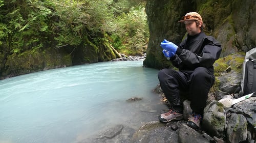 Researcher sits on a rock alongside a remote a bright blue-turquoise river mouth in olympic national park holding the Discovery eDNA surface sampler