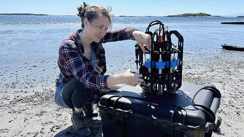 Woman kneels on the shore of a beach with her hands on Ascension eDNA depth sampler which rests on a black case on the sand