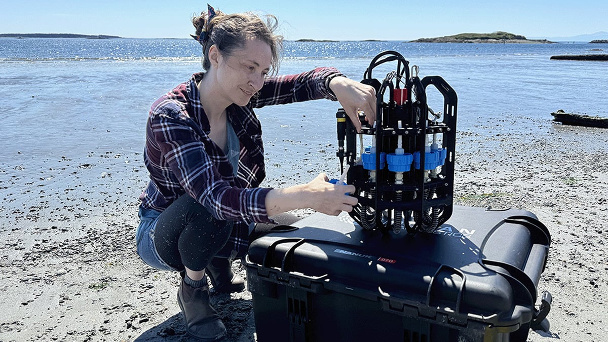 Woman scientist kneels on a sandy beach shoreline to look at the Ascension eDNA depth sampler sitting on a black carrying case.