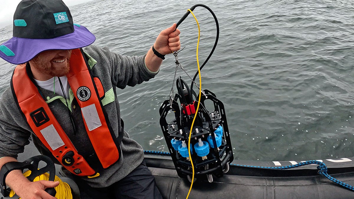 A researcher sits on the side of a small dinghy, holding a microplastics depth sampler in one hand.