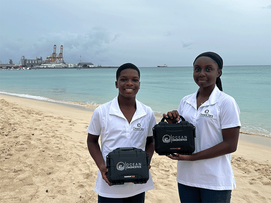 Two children stand on a tropical beach holding two carrying cases of microplastics survey tools