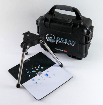 The Saturna Imaging System's tripod legs are placed overtop of a background sheet containing multi-coloured microplastic particles. A black carrying case with Ocean Diagnostics' logo sits behind the set up.