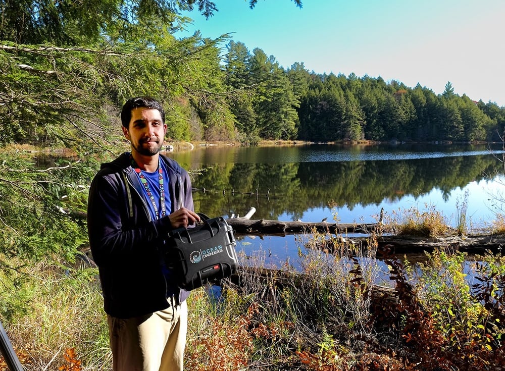 Researcher stands in front of a remote lake in Gatineau Park holding the eDNA Surface Sampler carrying case.