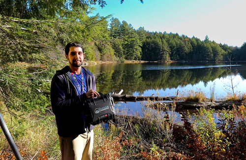 Researcher stands beside a remote lake in Gatineau Park holding up a small black carrying case that contains the eDNA Surface Sampler.