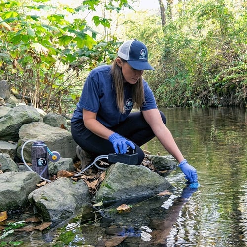 Scientist in a navy blue t-shirt wears latex gloves and holds a small portable black eDNA surface sampler resting on a rock as she collects eDNA from a stream.