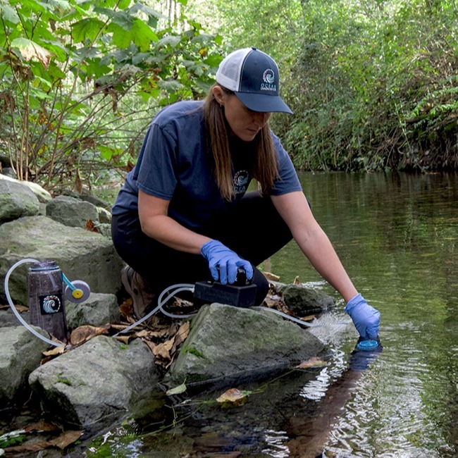 Woman kneels at a rock on the edge of a river creek surrounded by greenery collecting eDNA samplers with the portable hand-held sampler
