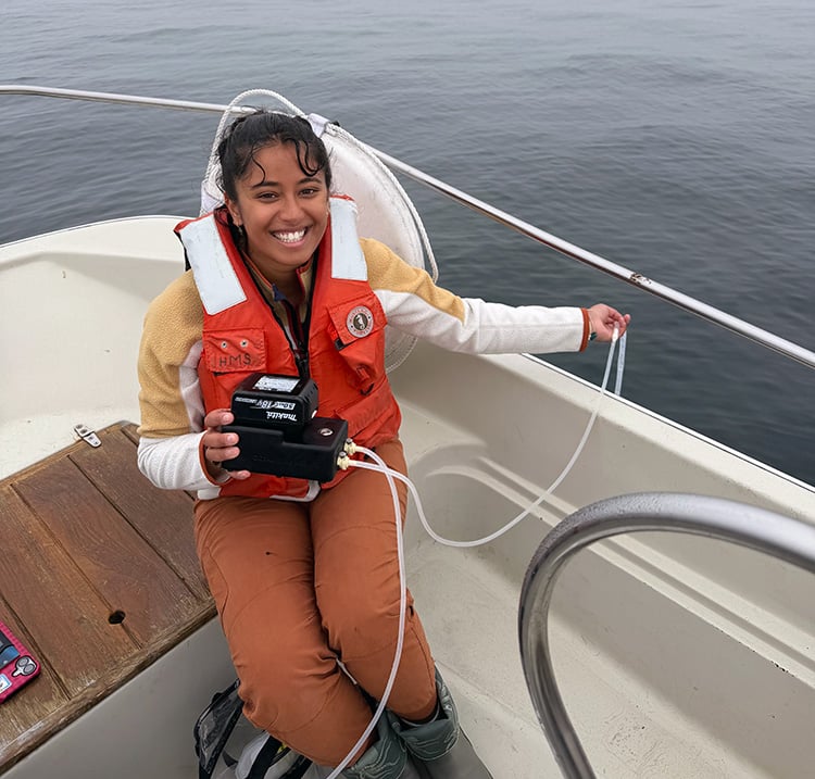 PhD student sits on the bow of a small boat smiling while holding the eDNA surface sampler in one hand and a tube that sits in the water from the side of the boat in the other