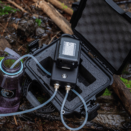 The hand-held eDNA Surface Sampler sits on top of its open black carrying case at a forest stream