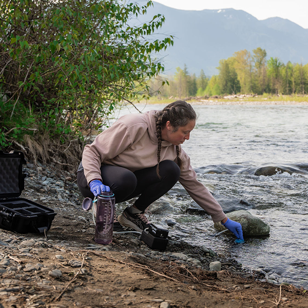 Woman kneels along the side of a river collecting eDNA samples with one hand while the surface sampler sits beside her foot