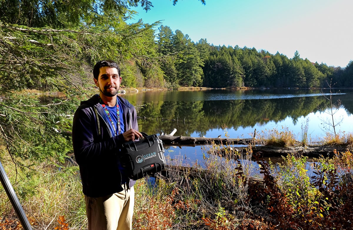 Researcher stands beside a remote lake in Gatineau Park holding up a small black carrying case that contains the eDNA Surface Sampler.