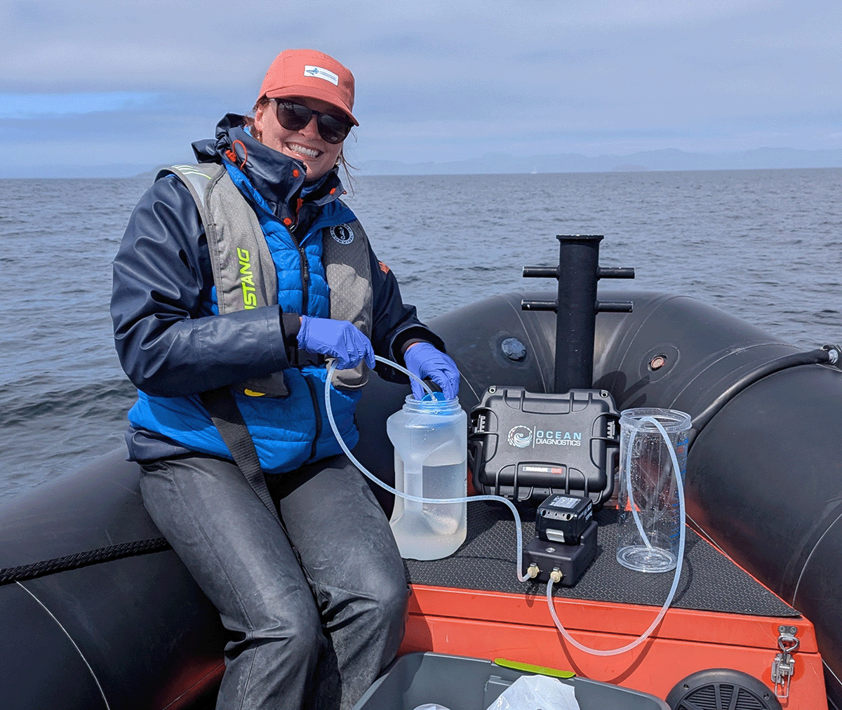 PhD researcher sits at the bow of a dingy in the ocean smiling and holding a filter into a jar of water as the eDNA surface sampler pump filters water and collects eDNA on her filter.