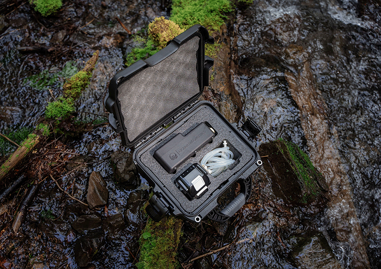 A waterproof black case is open and sits on a log in a forest creek. From above, foam cutouts house the eDNA surfaace sampler, a battery and tubing. 
